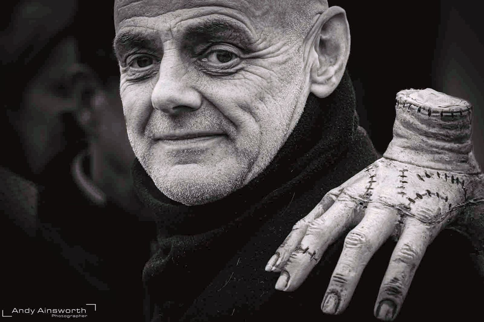 Older man in Addams Family costume with a stitched-hand prop on his shoulder, black and white — Whitby Goth Weekend