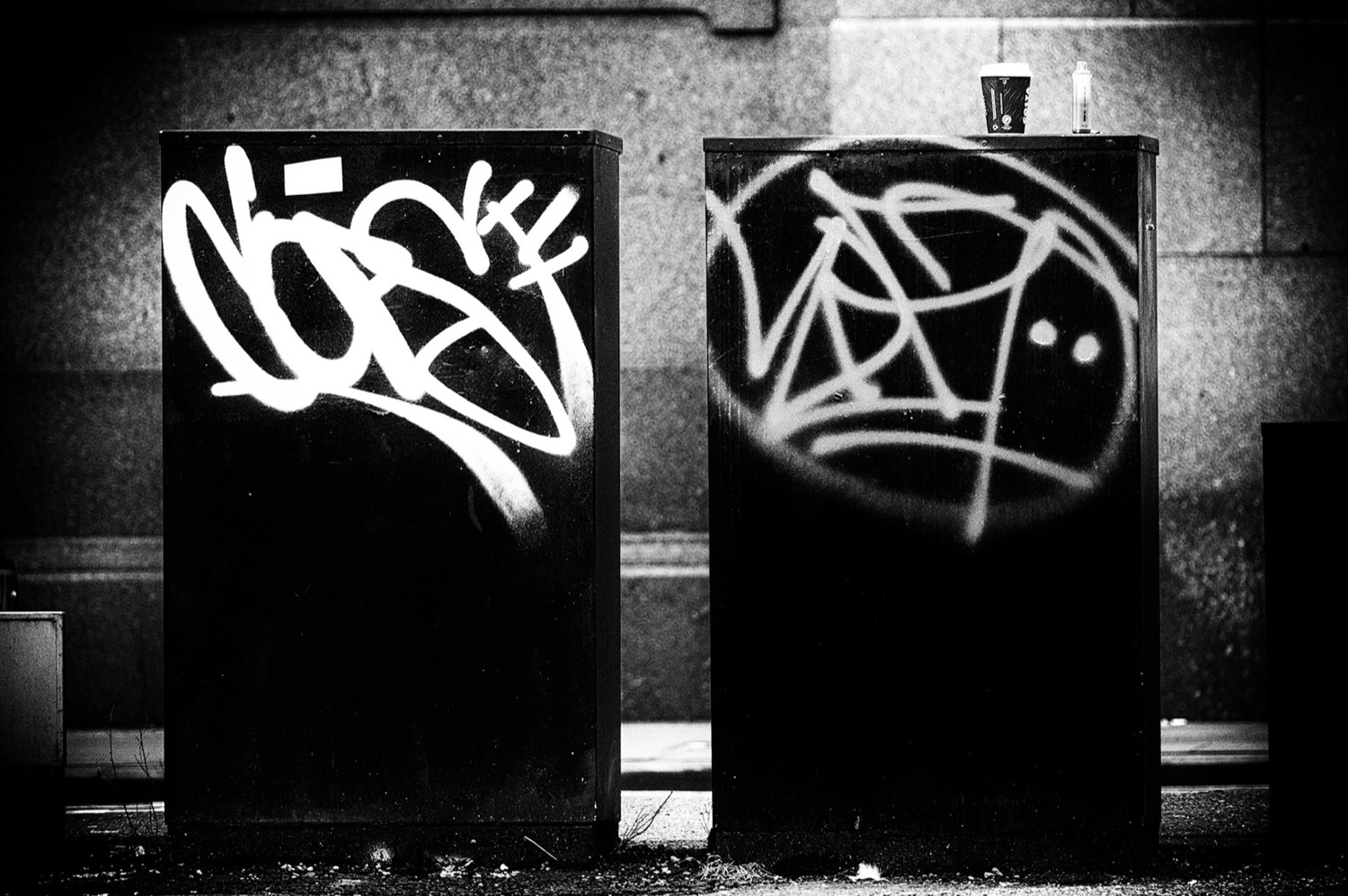 Two tagged black utility boxes, one spray-painted tag forming an accidental face — street photography, Manchester